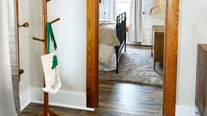 A room with a wooden coat rack, a bed with a white comforter, a wooden floor, and a doorway leading to another room. - Sunny Downtown Gunnison Retreat (Gunnison)