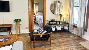 A cozy living room with a wooden floor, a black leather sofa, a round wooden coffee table, a small wooden side table with a plant, a TV mounted on the wall, a round mirror, and a window with curtains. - Sunny Downtown Gunnison Retreat (Gunnison)
