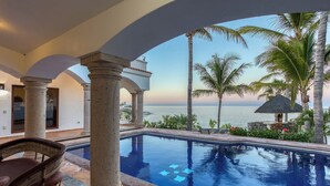 A pool area with a view of the ocean, surrounded by palm trees and a thatched umbrella.