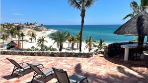 A patio with outdoor seating, a thatched umbrella, and a view of a beach with palm trees and the ocean.