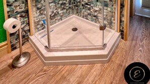 Ducha con mampara de vidrio, pared de mosaicos de piedra, suelo de madera y un dispensador de papel higiénico visible.