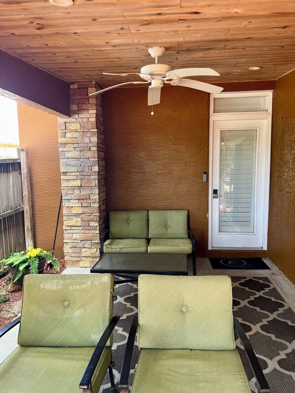 Un patio techado con ventilador de techo, pared de piedra, puerta con panel de vidrio y zona de estar con dos sillones y una mesa.