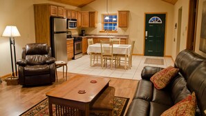 A cozy living room with a leather sofa, a wooden coffee table, and a dining area with a table and chairs. A kitchen with wooden cabinets and appliances is visible in the background. - Hot Tub Cabin- In the Heart of Shawnee Wine Trail (Carbondale)