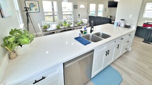 Et moderne køkken med hvid bordplade, stålvask og sorte skabe. Der er hængende pendellamper, et spiseområde med et bord dækket til fire personer, og et stort vindue der giver naturligt lys.