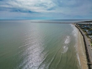 An aerial view of a coastal area with a beach, a breakwater, and a row of houses along the shore.
