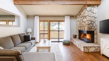 Cozy living room featuring a large, comfortable sofa, high ceilings, and a beautiful stone fireplace—perfect for relaxing after a day on the mountain. Step out onto the private balcony and enjoy incredible views where you can actually watch skiers coming