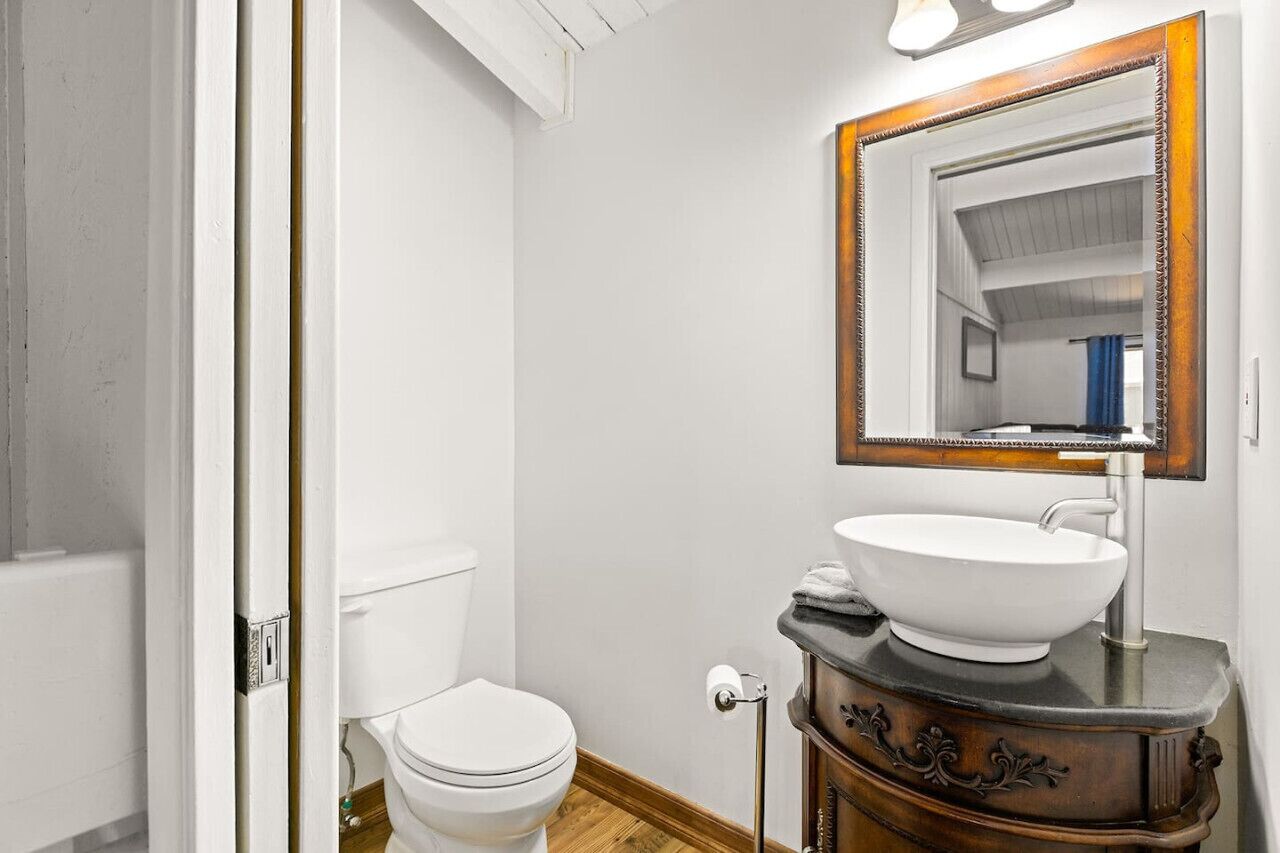 Un baño con inodoro blanco, tocador de madera con lavamanos blanco y espejo con marco de madera.