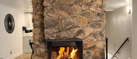 Una chimenea de piedra con fuego, rodeada por un banco de madera y una mesita con una planta.
