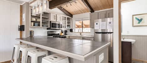 A modern kitchen with a large island, bar stools, stainless steel appliances, and a wooden ceiling.