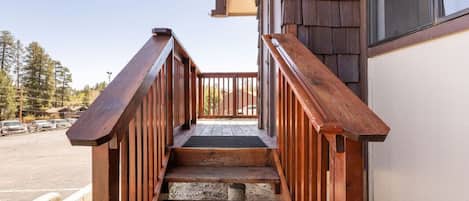 A wooden staircase leading to a porch with a view of a parking lot and trees.
