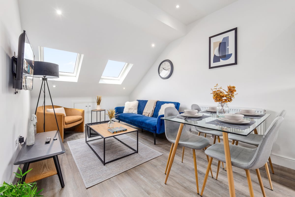 A modern living room with a sofa, coffee table, and dining table. There are skylights and a clock on the wall.
