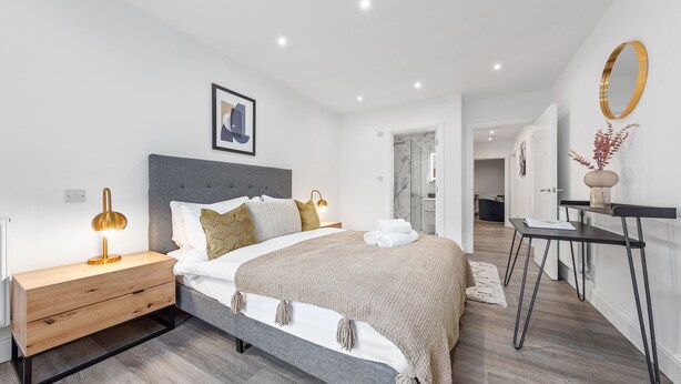 Ein modernes Schlafzimmer mit Bett, Nachttisch und Nachtlicht. An der Wand hängt ein Spiegel und eine Tür führt in einen weiteren Raum.