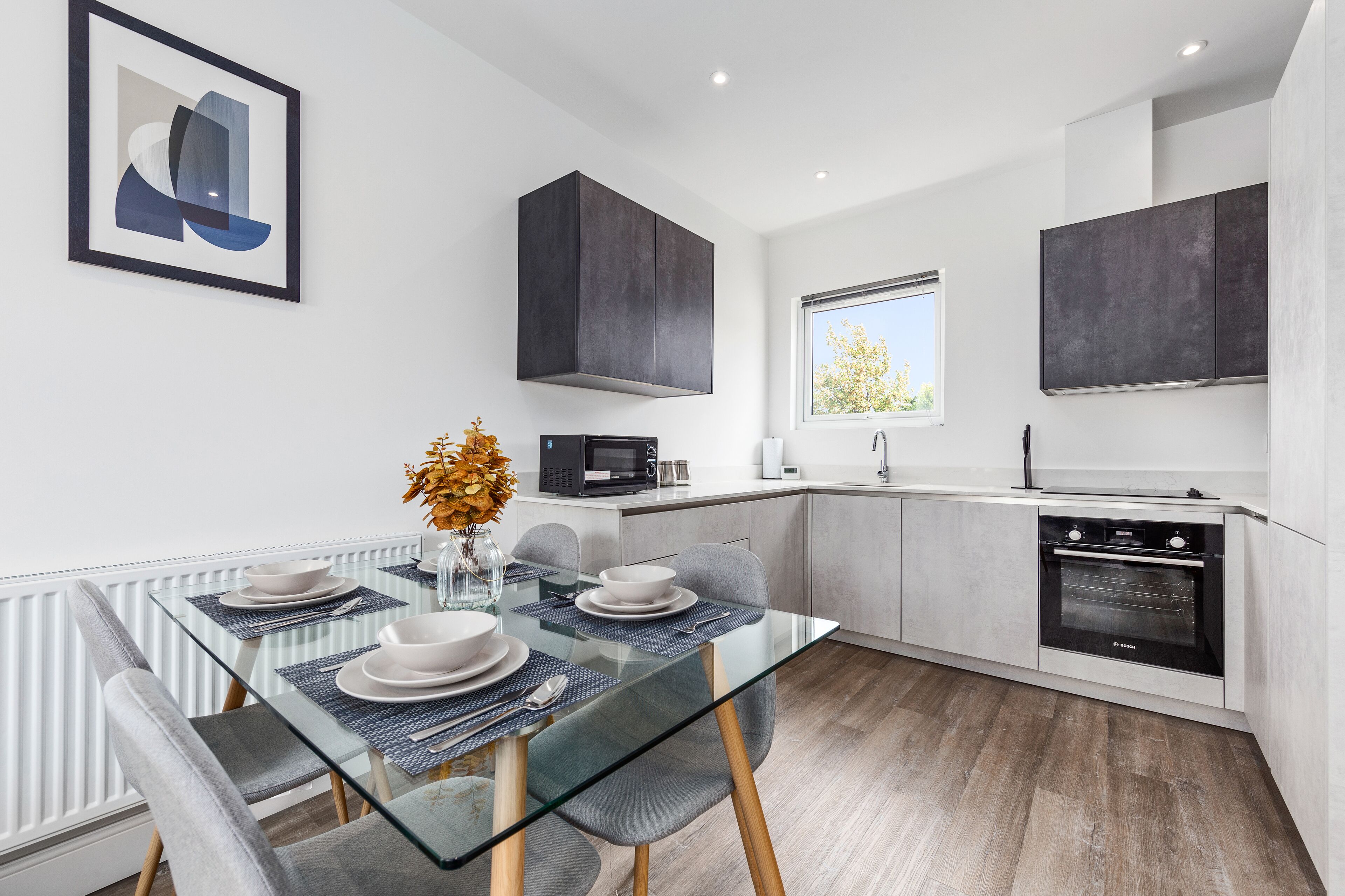 A modern kitchen with a glass dining table, grey chairs, and a Bosch oven.