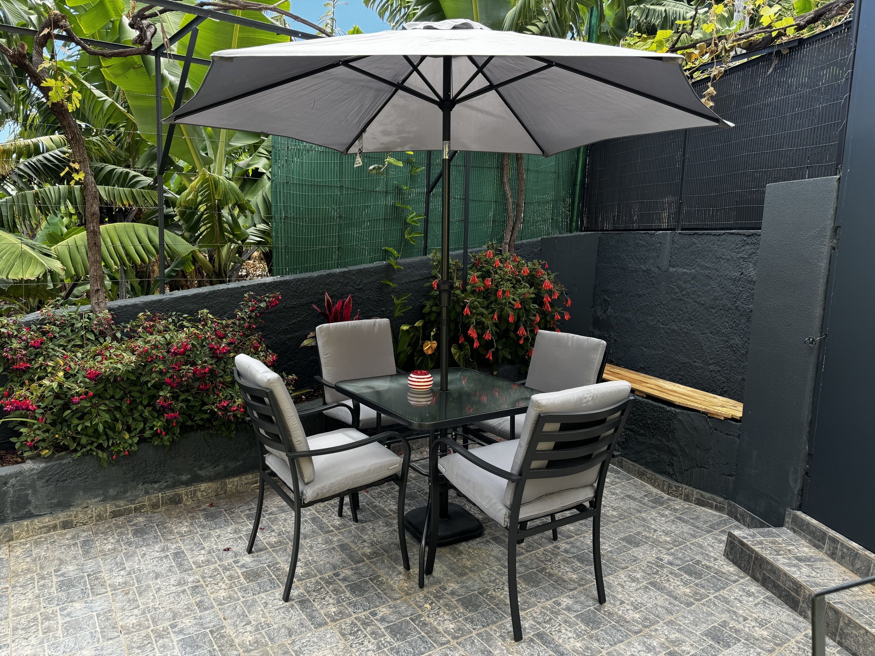 Une terrasse avec une table et des chaises, un parasol et un mur noir agrémenté d’une clôture verte.