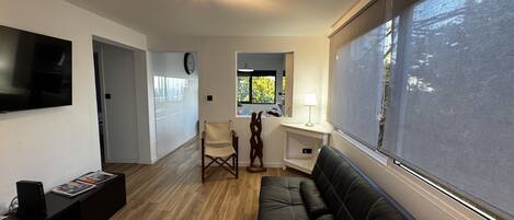 A modern living room with a black leather sofa, a wooden floor, a flat-screen TV mounted on the wall, a small table with a lamp, and a window with a view of greenery.