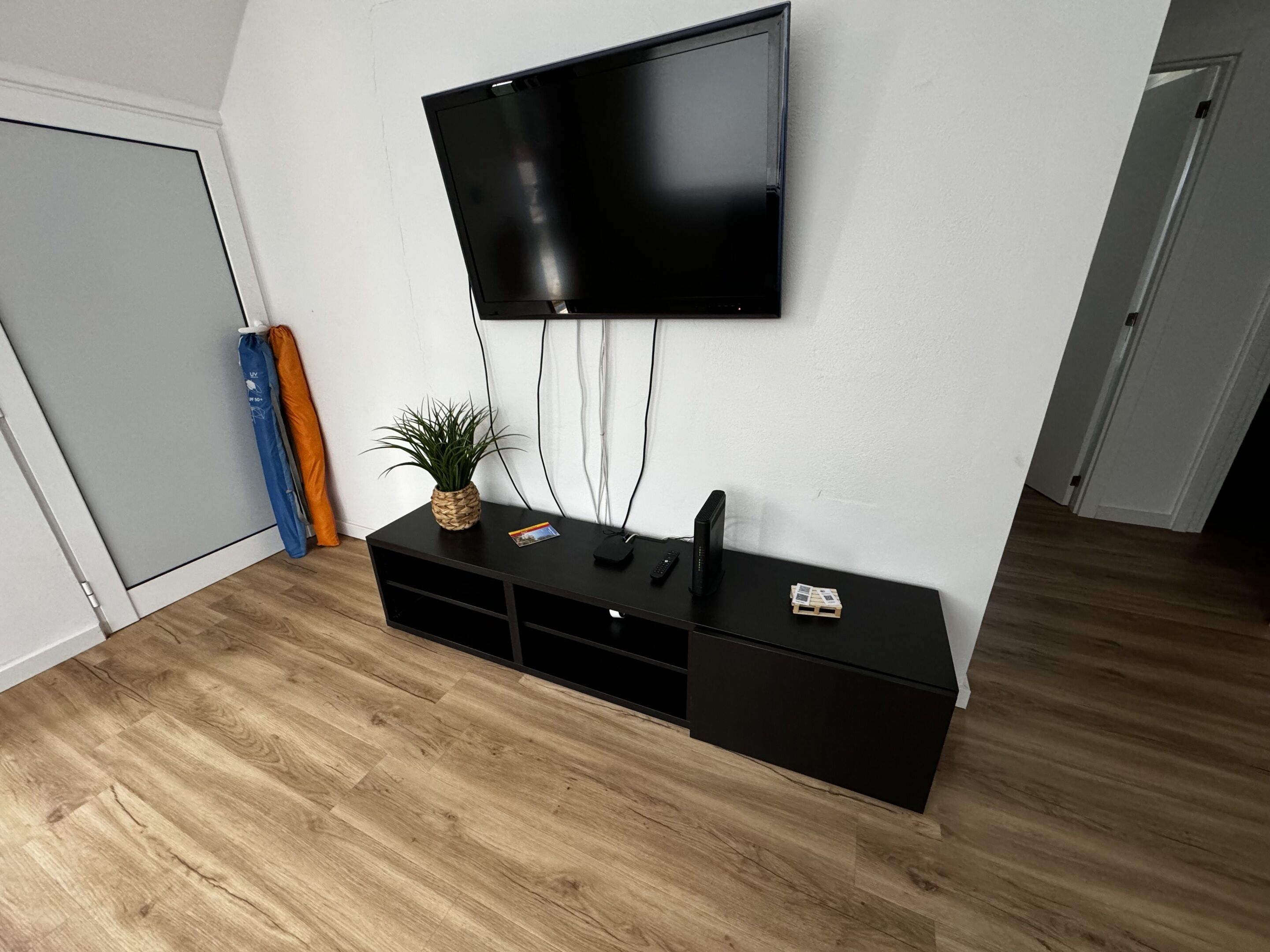 A modern living room with a flat-screen TV mounted on the wall, a black entertainment unit with a plant, and a wooden floor.