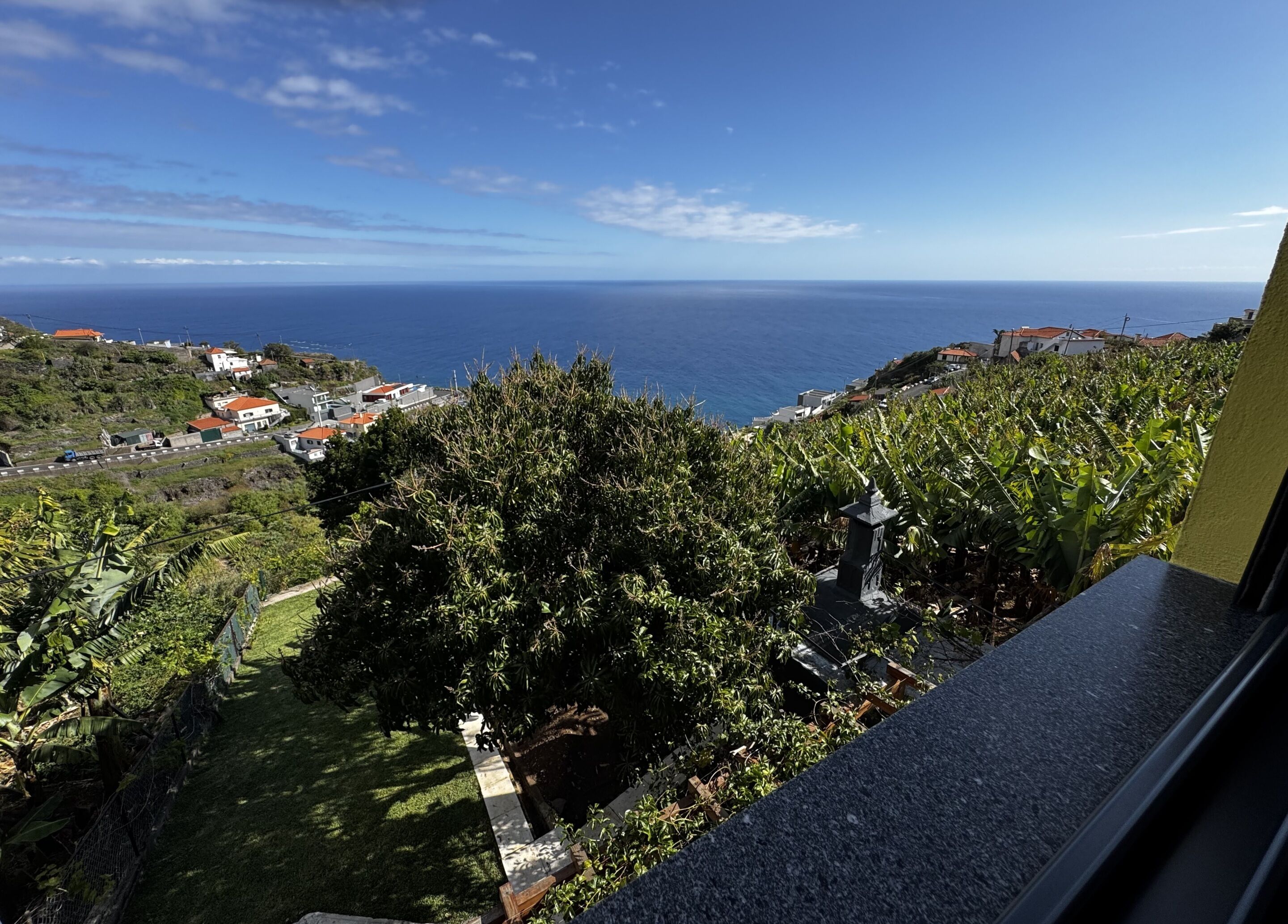 A coastal view from a balcony overlooking a garden with a stone pathway, a small pond, and a view of the sea and distant buildings.