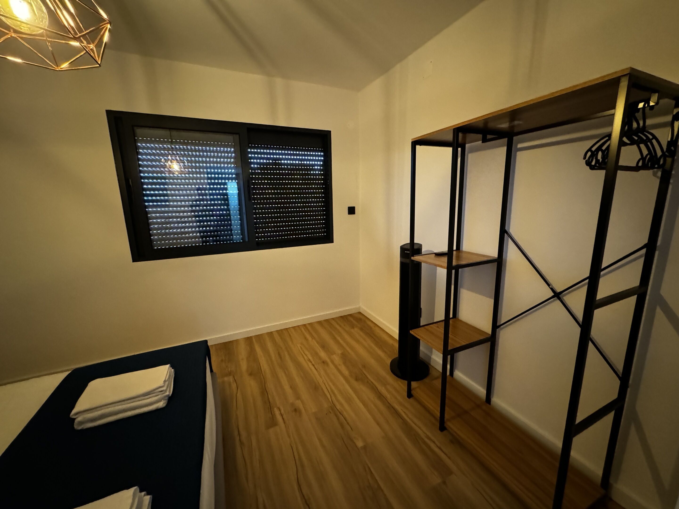 Ein Zimmer mit Fenster, Holzboden, einem schwarzen Metallregal und einem kleinen Tisch mit weißem Tuch.
