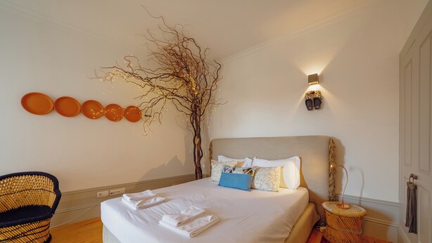 A neatly made bed with white linens and a blue pillow, a wicker chair, and a wall-mounted light fixture. - Downtown Modern Studio by Vacationy (Porto)