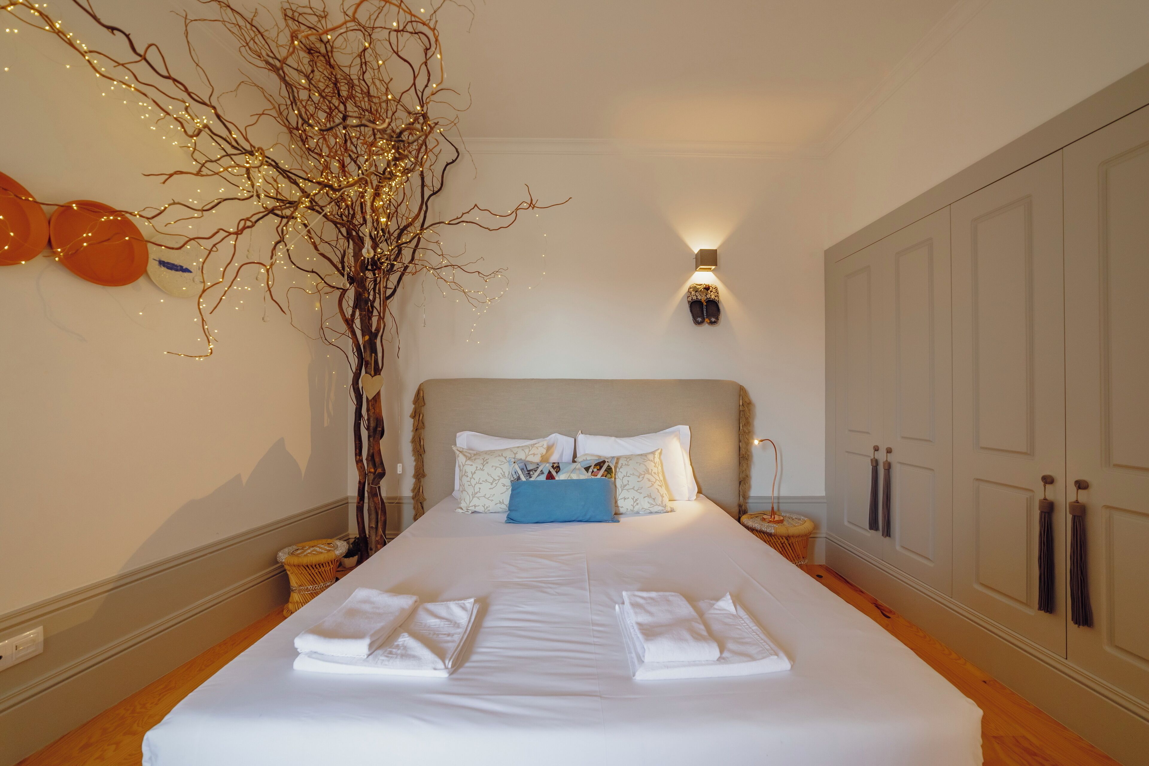 A neatly made bed with white linens, a blue accent pillow, and a decorative branch with lights.