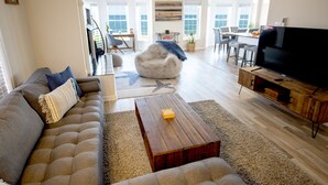 A spacious living room with a sofa, coffee table, and TV. There is a dining area with chairs and a table, and a view of the ocean through large windows. - Beachfront Getaway, pet friendly (Norfolk)