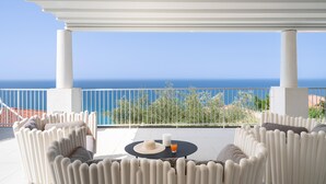Ein überdachter Außenbereich mit weißen Stühlen und einem Tisch, mit Blick auf eine schöne Aussicht auf das Meer und viel Grün.