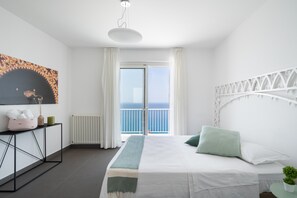A modern bedroom with a large bed, a bedside table, a radiator, a window with curtains, and a view of the sea.