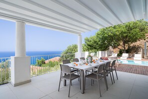 A covered outdoor dining area with a view of the sea, a pool, and a tree.