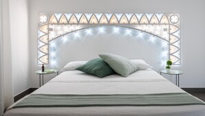Ein ordentlich bezogenes Bett mit weißer und grüner Tagesdecke, zwei Kissen und einer grünen Decke. Neben dem Bett steht ein kleiner runder Tisch mit einer Pflanze darauf.