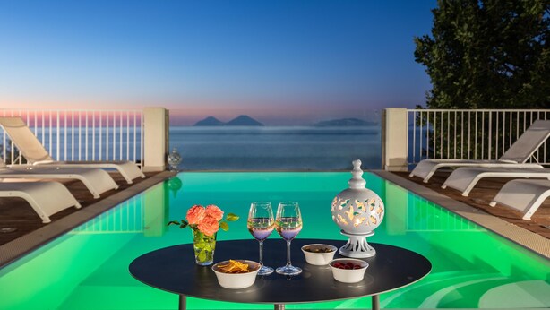 Ein Tisch am Pool mit Getränken und Snacks, Liegestühle und Blick auf Meer und Berge.