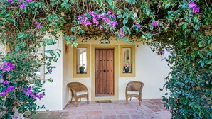 A building with a wooden door, two wicker chairs, and purple flowers. - Calle del Marmol 162 (Manilva)