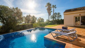 A swimming pool with lounge chairs, surrounded by a garden and a building with a tiled roof. - Calle del Marmol 162 (Manilva)