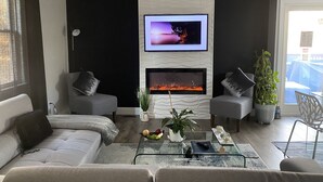 A modern living room with a flat-screen TV mounted above a fireplace, two grey sofas with cushions, a glass coffee table with decorative items, and a potted plant. - Contemporary Cozy Cottage (Lake Peekskill)