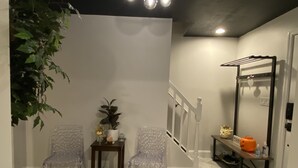 A room with a white wall, a wooden staircase, a clear plastic chair, a small wooden table with a plant, and a metal rack with a pumpkin. - Contemporary Cozy Cottage (Lake Peekskill)