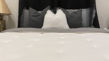 Cama com edredom branco, almofadas cinzas e cabeceira preta.