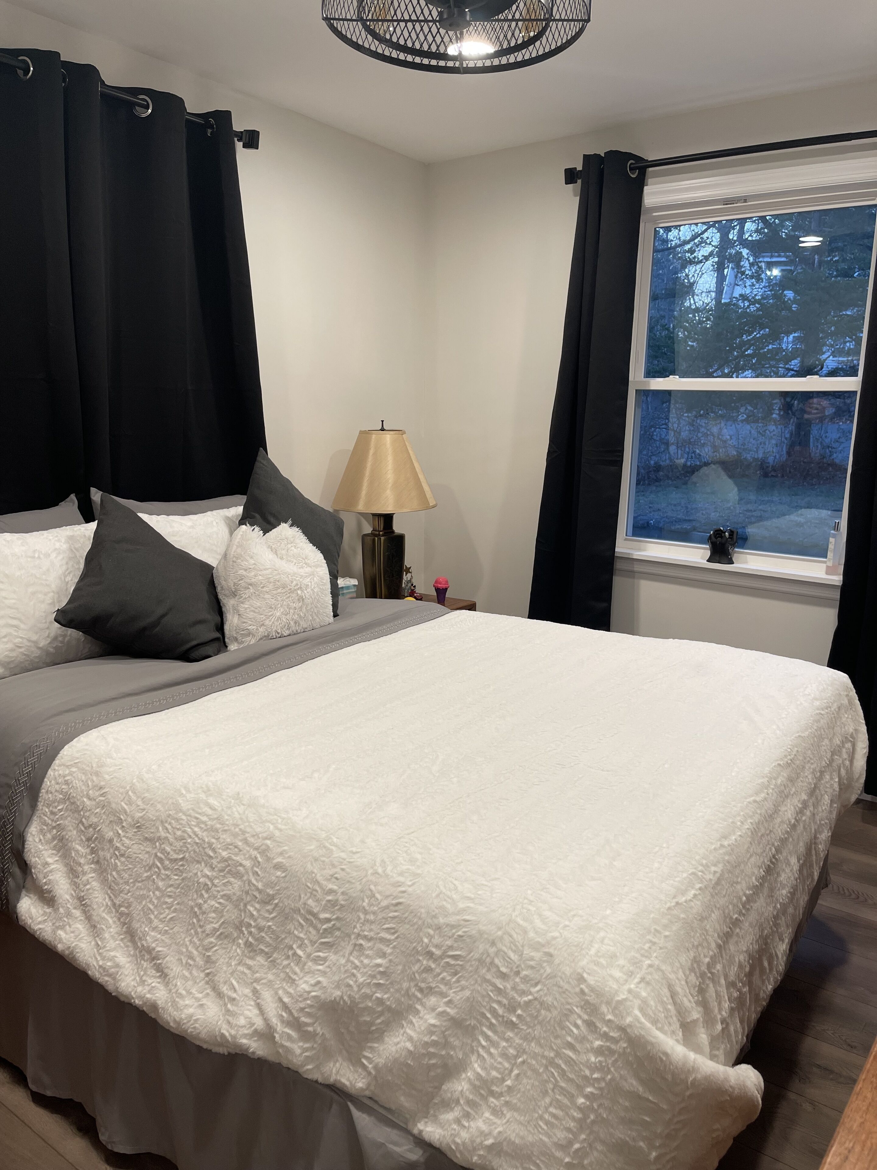 A bedroom with a large bed, a bedside table with a lamp, and a window with a view of trees.