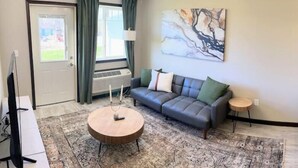 A living room with a grey sofa, a round coffee table, a painting on the wall, and a window with curtains.