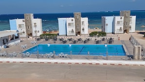 Una piscina con múltiples carriles, rodeada de sillones y sombrillas, con vista al mar y edificios al fondo.