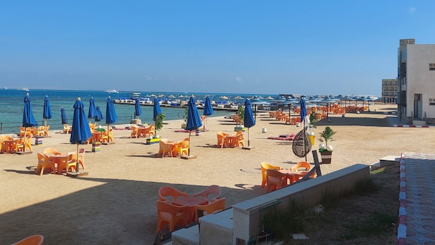Una playa con hileras de sillas de plástico naranja y sombrillas azules, una zona arenosa y un edificio al fondo.