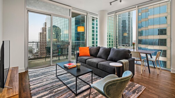 Una sala de estar moderna con sofá, mesa de centro y sillas. Cuenta con ventanas amplias con vista a la ciudad y un balcón.