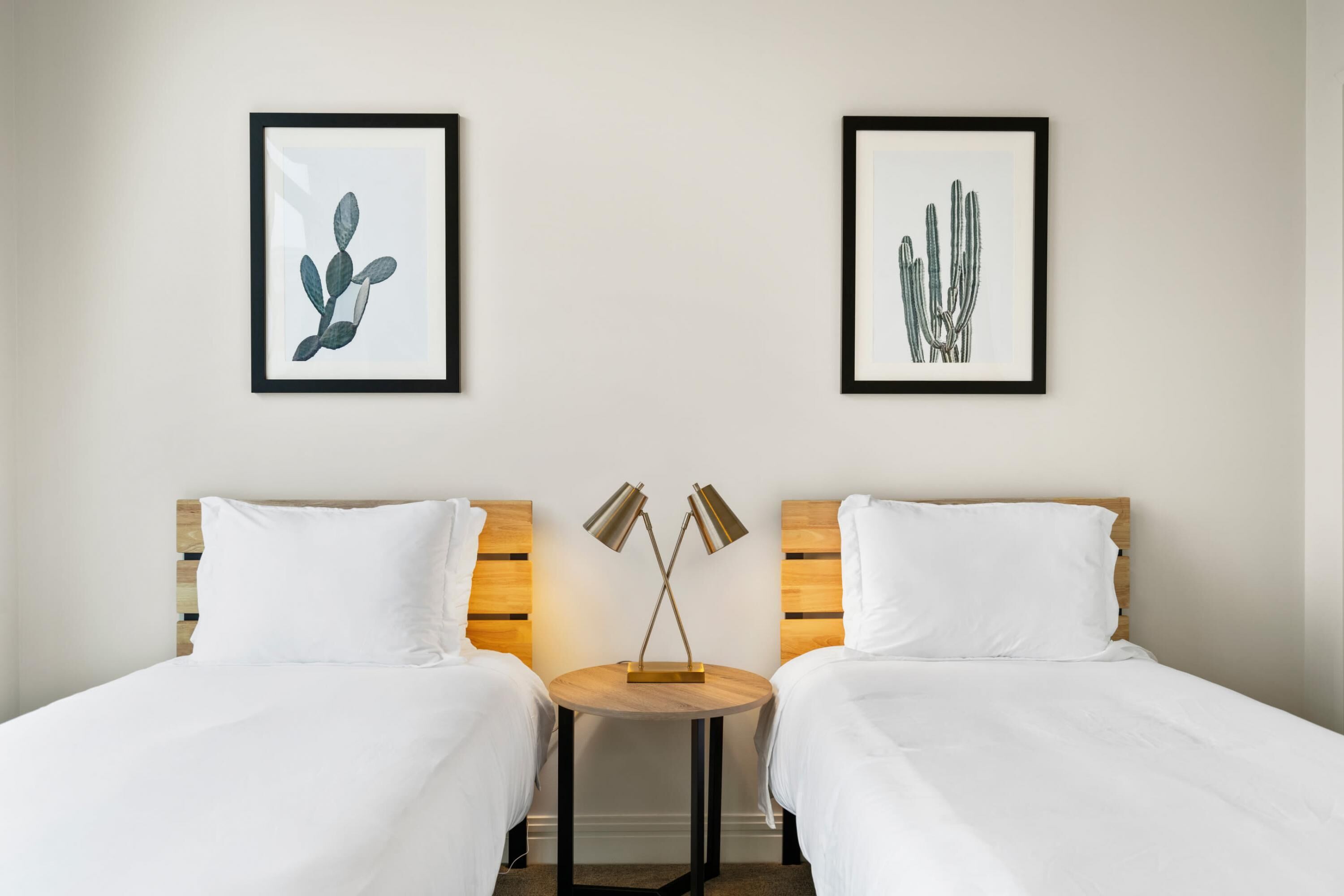 Ein Hotelzimmer mit zwei Betten, einem runden Tisch mit zwei Lampen und gerahmten Pflanzenbildern an der Wand.