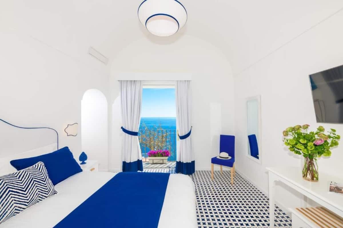 A hotel room with a bed, a chair, a TV, and a view of the sea.