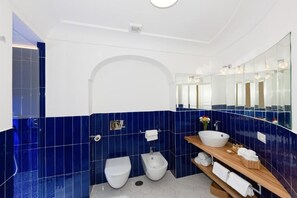 Un baño moderno con azulejos azules, un inodoro blanco, un bidé, un mueble de tocador de madera con un lavabo y un espejo grande.
