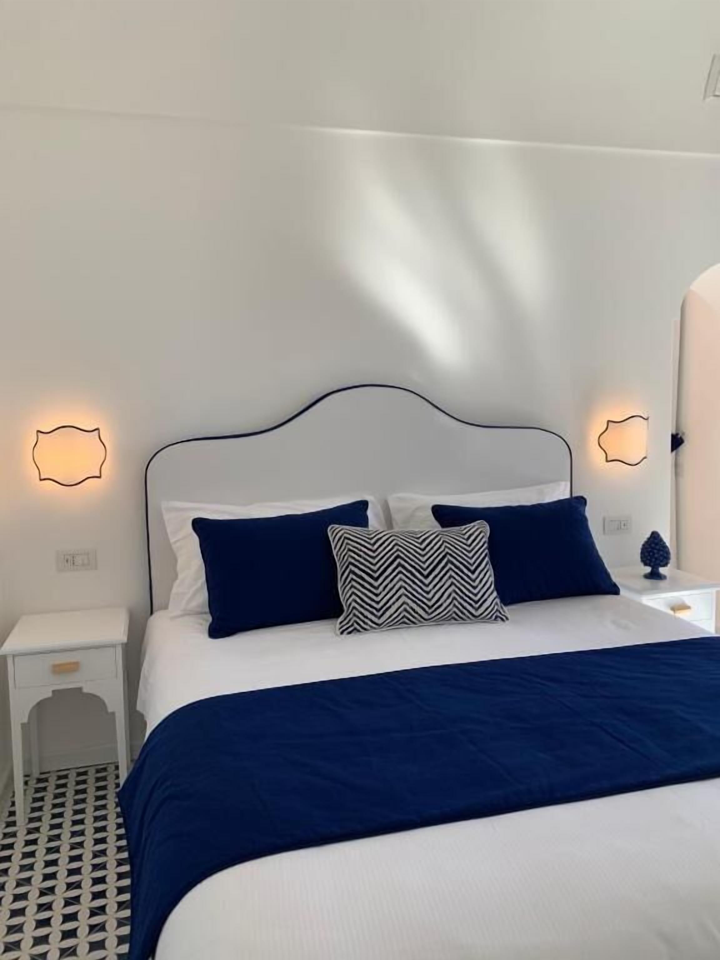 A neatly made bed with a white headboard and navy blue bedding. There are two bedside tables, each with a small lamp. The room has a clean, modern design with white walls and a geometric patterned floor.