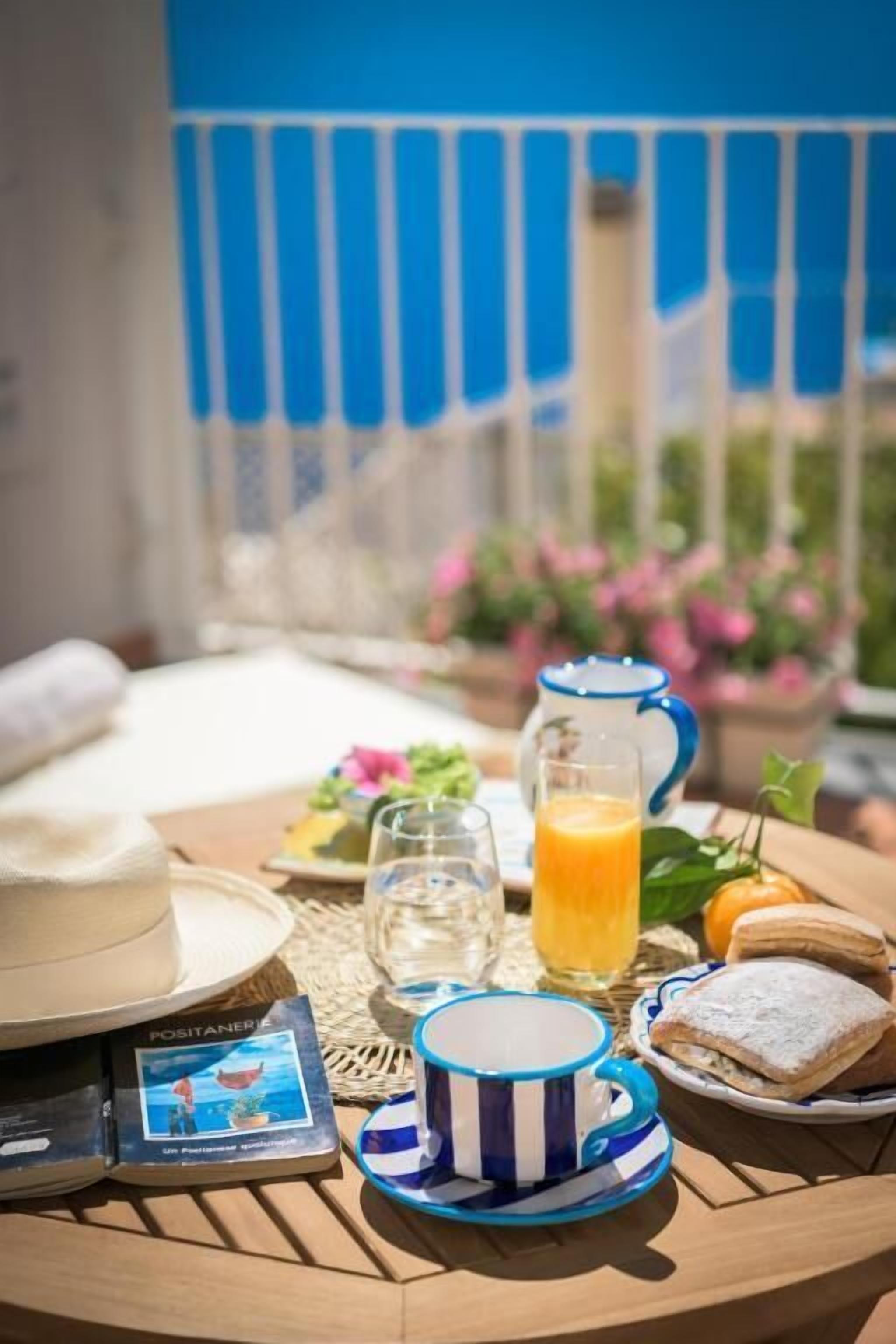 Una mesa con un libro, una taza a rayas, un vaso de jugo de naranja, una jarra y un plato con comida en un balcón.