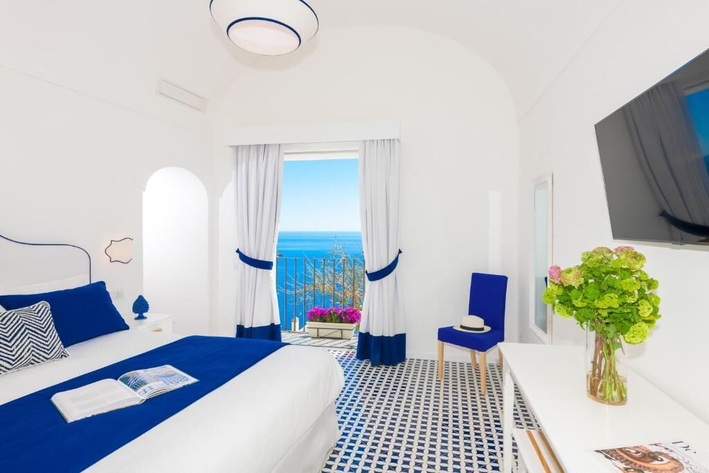 A hotel room with a bed, a small table with a vase of flowers, a chair, and a view of the sea.