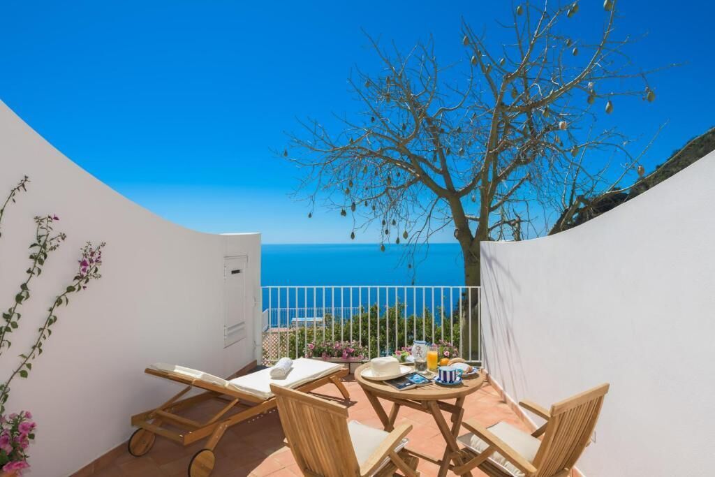 Ein Balkon mit Meerblick, Holzstühle, ein Tisch mit Buch und Getränken, und ein Baum.