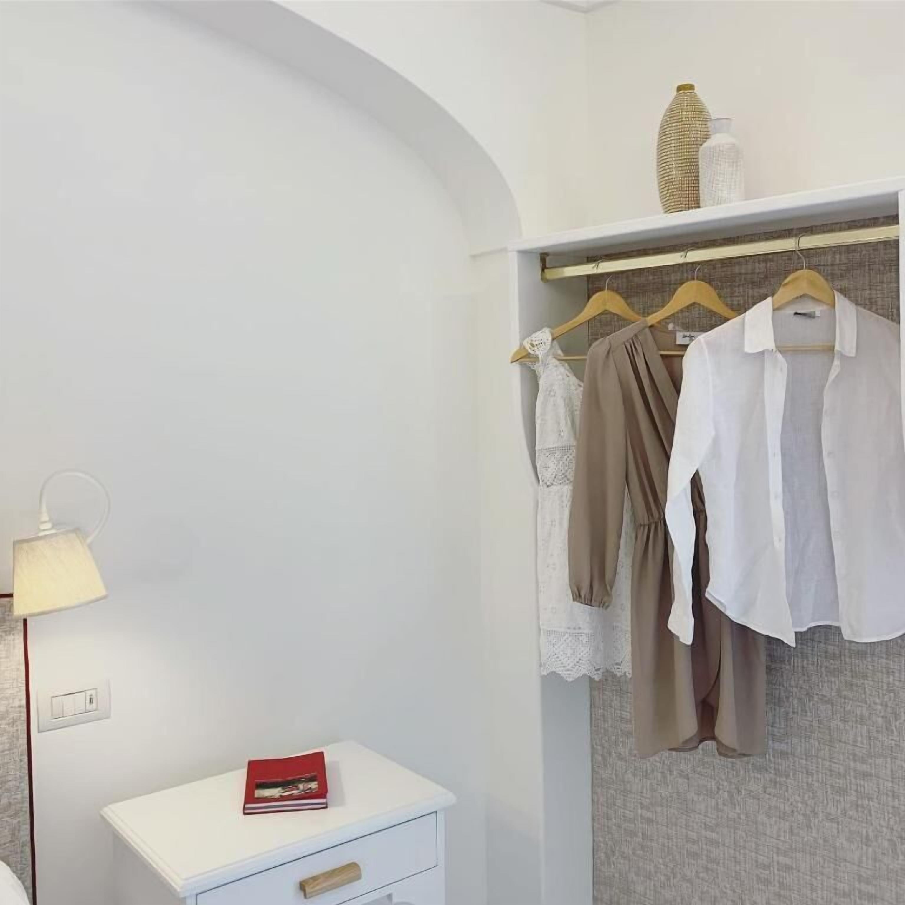 Quarto com cômoda branca, abajur e guarda-roupa com roupas.