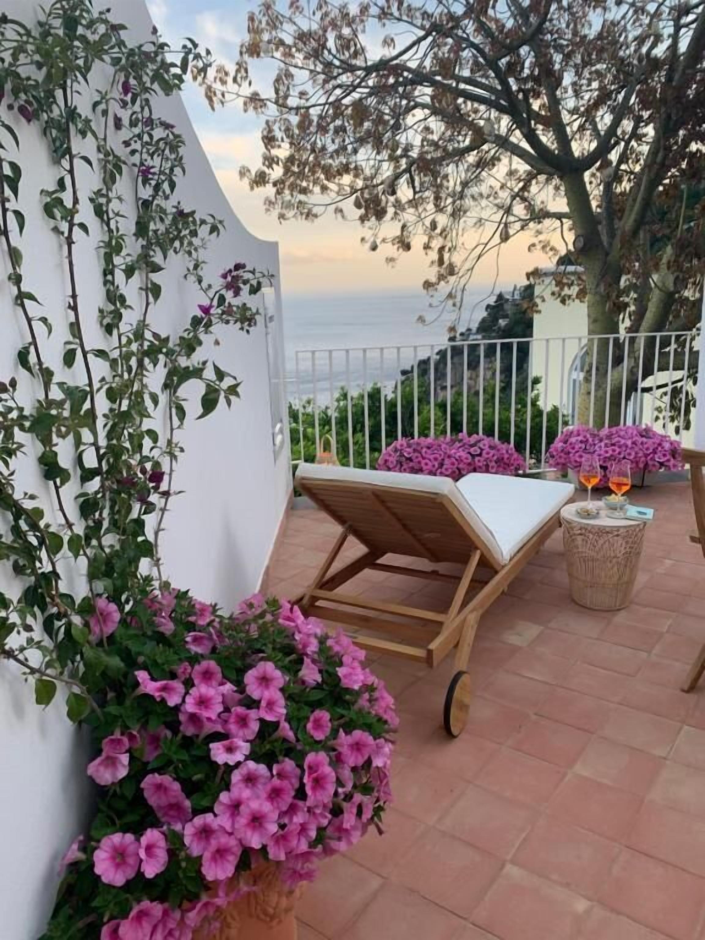 Un balcón con una silla de lounge de madera, una mesita con bebidas y macetas con flores rosadas.