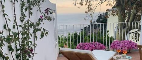 Un balcón con una silla de lounge de madera, una mesita con bebidas y macetas con flores rosadas.